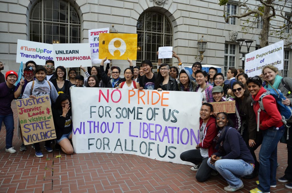 Image description: A large crowd of APIENC members and supporters are standing outdoors holding signs and a big banner that says: "No Pride for Some Of Us Without Liberation For All Of Us."