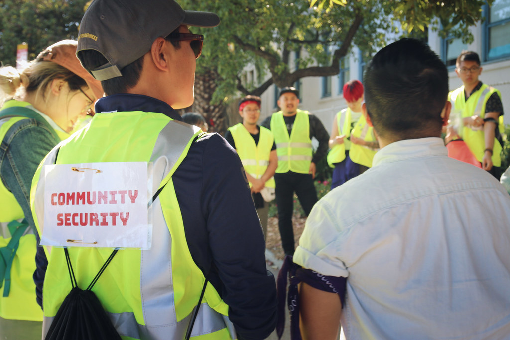Image description: a group of TGNC API people and allies stand outdoors in a circle. Most are wearing bright yellow safety vests and say "Community Security" on the back.