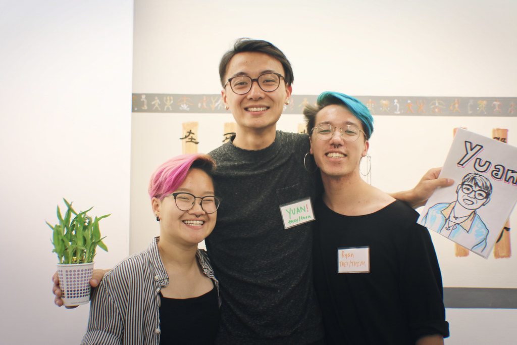 Image description: Two summer organizers (Huanvy, Rai) are standing next to community organizer (Yuan) and smiling indoors. Yuan is holding a green plant and a drawing of themself.