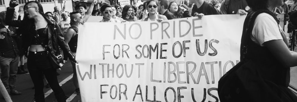 Image description: A black and white photo of the API contingent at 2015 SF Trans March. They are holding a big banner that says: "No Pride For Some Of Us Without Liberation For All Of Us."