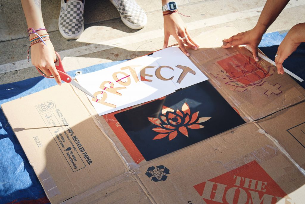 Image description: There are two pairs of hands laying stencils down of the word "PROTECT" and an image of a lotus flower on a piece of cardboard.