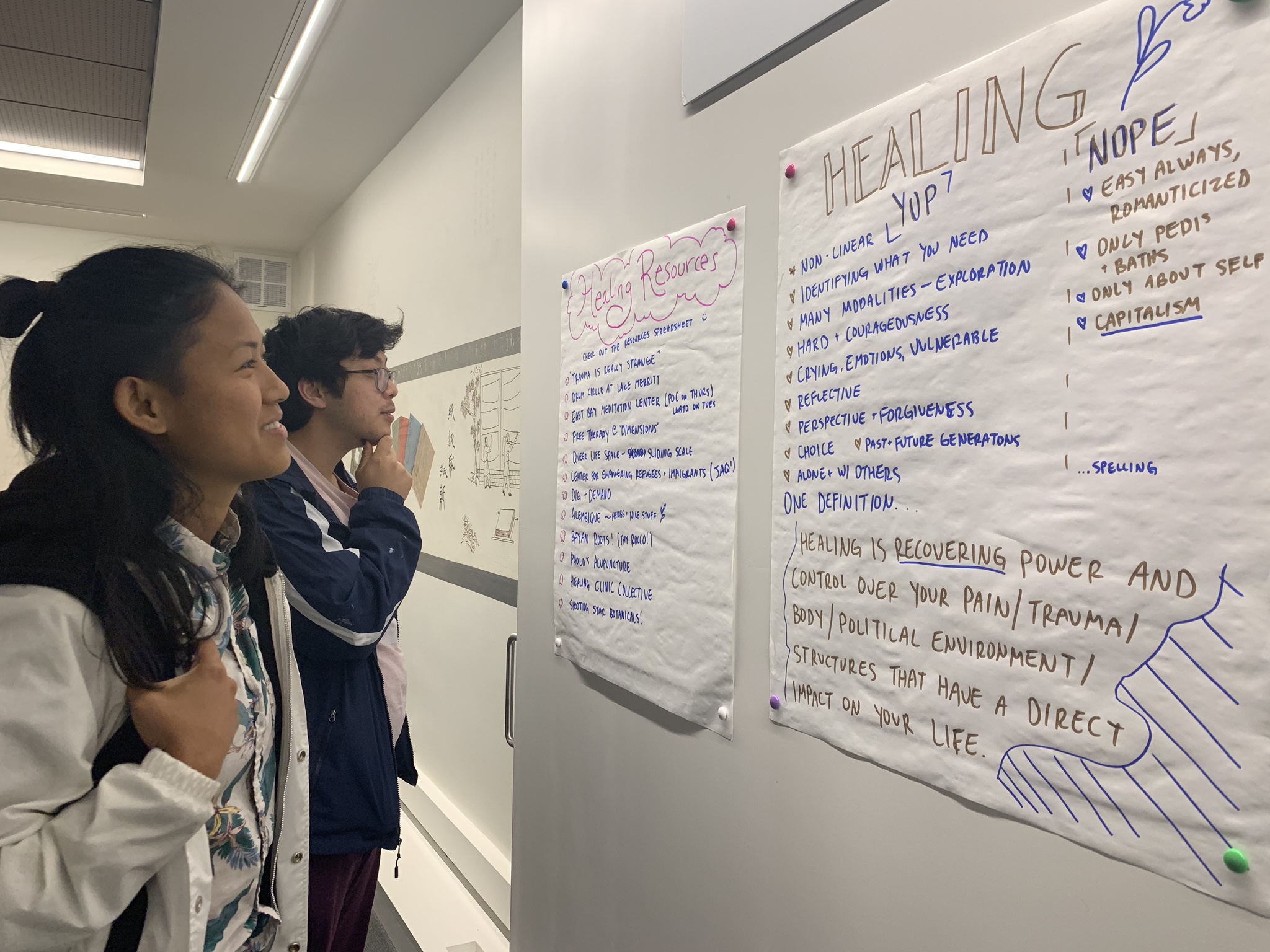 Image description: Two APIENC members are indoors looking at flipchart papers attached to the wall. They are about healing and healing resources.