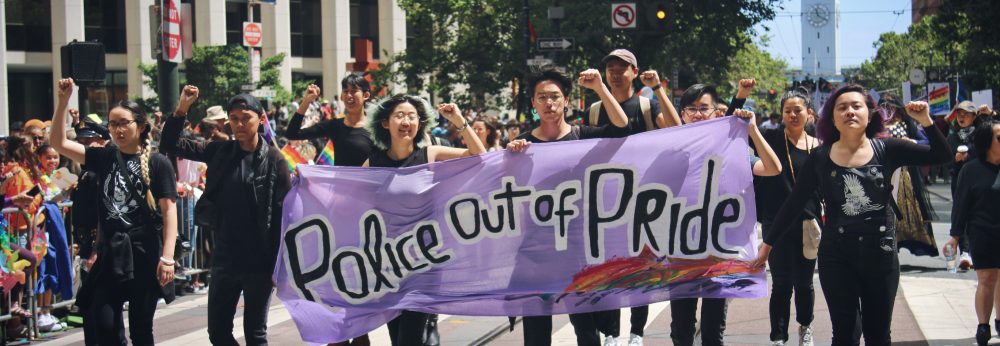 Image description: APIENC members are dressed in black with fists raised marching down Market St. at SF Pride Parade 2019. They are holding a purple banner that reads: "Police out of Pride."