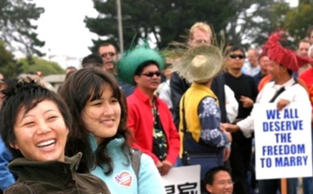 Image description: Two TQAPI people are in the foreground smiling. There are several QTAPI and allies in the background holding signs like "We all deserve the freedom to marry."