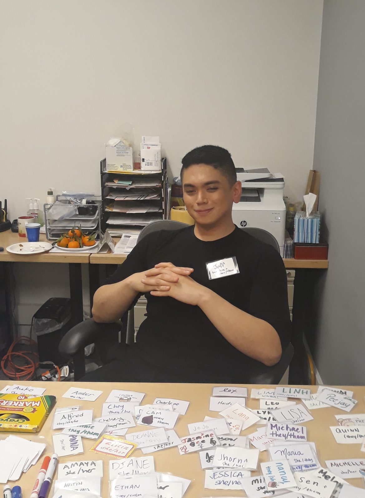 Image description: Jeff is sitting behind a desk indoors, smiling with hands folded in front of them. There are reusable name tags in front of them.