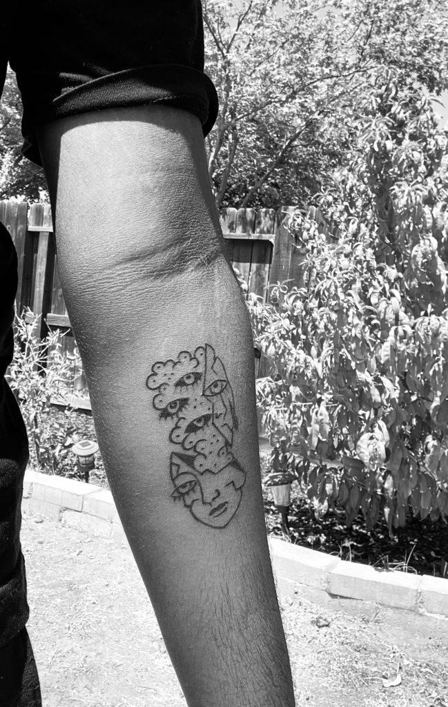 Image description: A black & white photo of Sajneel's right forearm. There is an abstract tattoo of a person's head exploding and three bold eyes flying in different directions.