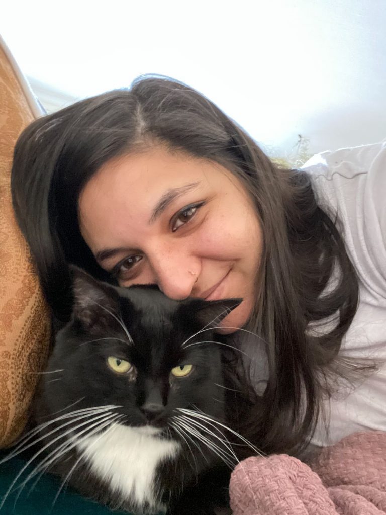 Image Description: Nisha leans onto an orange couch pillow over her cat, smiling at the camera. Her cat is black with a white patch, yellow eyes, and white whiskers.